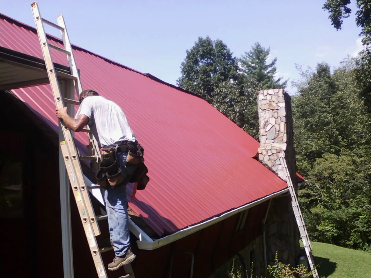 Roofer on a ladder beside a red metal roof and stone chimney — Soffit & Fascia Repair by Roofing Brothers in Belvue